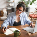 Woman working at a desk with a laptop and notebook in a home office setting.
